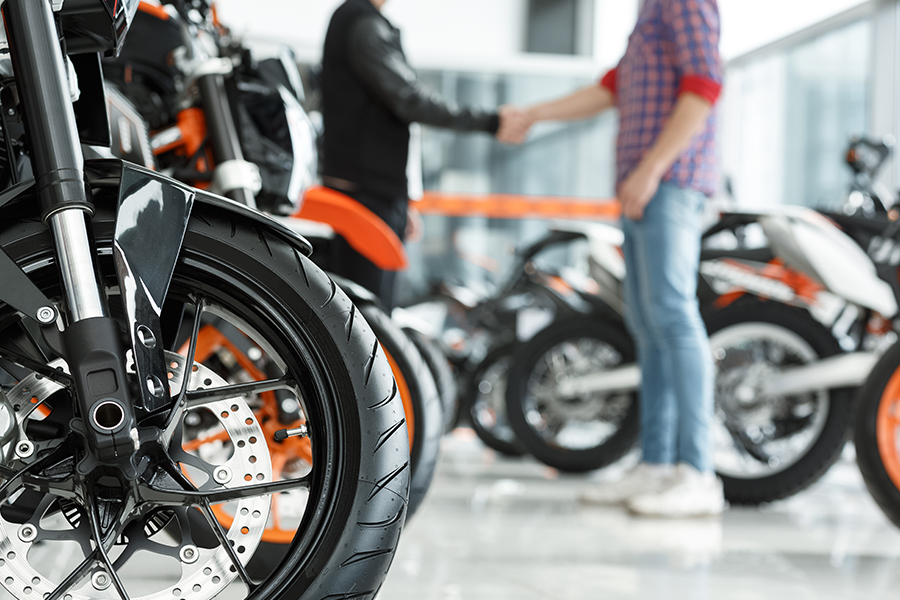 Motorcycle Dealership Insurance - Focus on a Motorcycle Wheel with Male Customer Shaking Hands with Motorcycle Dealership Manager in the Background