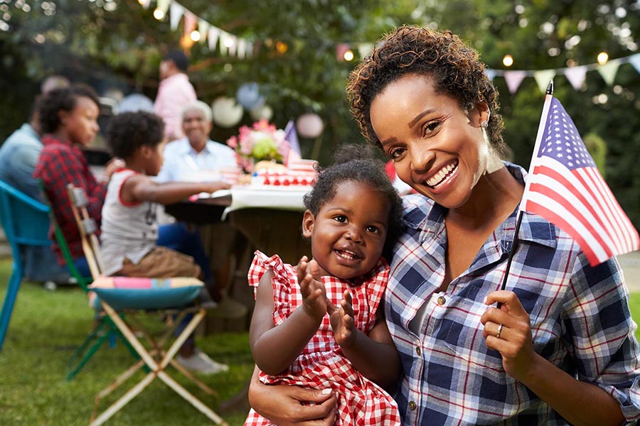 Blog - Mother and Daughter Hold a Small American Flag, Cheerful Summer Barbecue Behind Them on a Summer Day