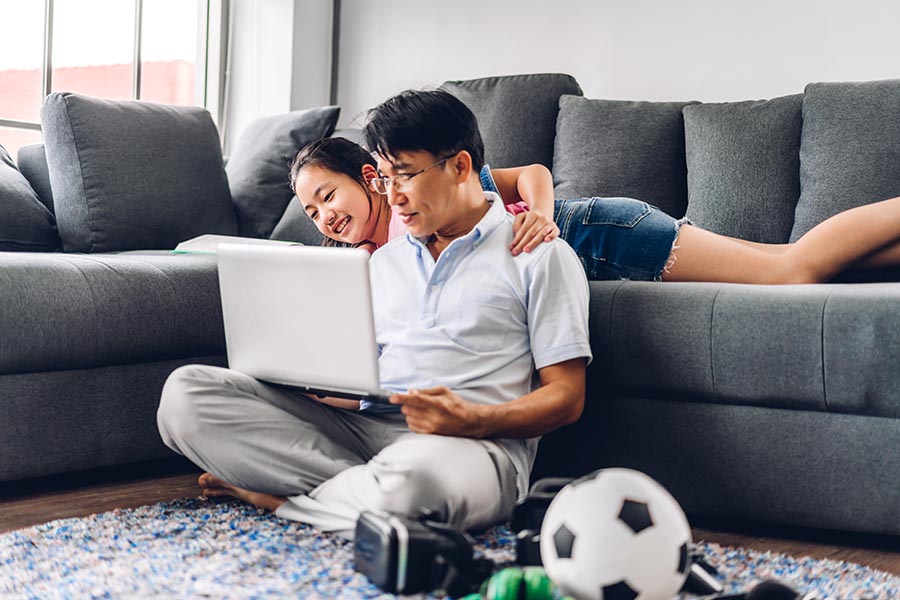 Client Center - Father Uses a Laptop on the Floor as His Daughter Watches From the Couch, a Soccer Ball Nearby