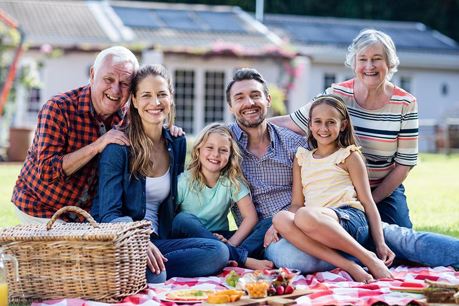 Personal Insurance - Three Generations Smile and Sit on a Picnic Blanket, a Picnic Basket and Plates of Food Spread Out, a Lovely Home Behind the Family